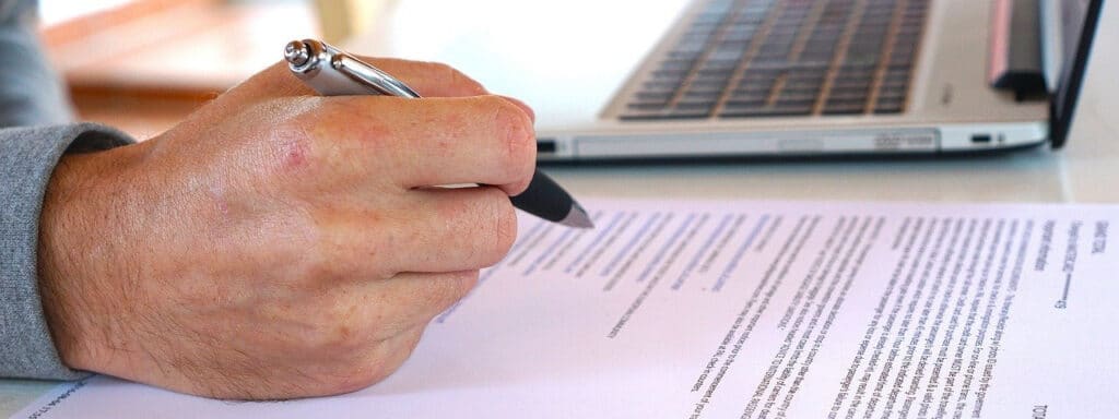 Main d'une personne âgée avec stylo et ordinateur relisant un contrat